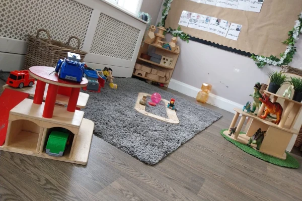 Wooden toy garage with red tabletop sits amid scattered toy cars and animal figures on a gray rug, inside a bright children's playroom with low shelves and a bulletin board.