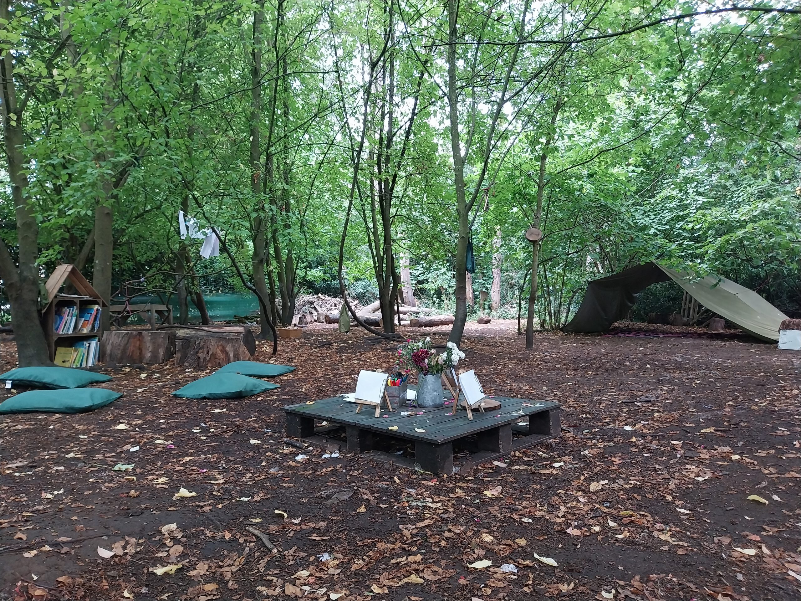 Wooden pallet holds small easels, art supplies and a vase of flowers; cushions and log seats sit nearby, forming a classroom in a leafy woodland clearing with tarp shelters.