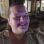 Woman smiling for a close-up selfie, wearing a sequinned top and necklace; lit by colored light. Background shows patio furniture and windows revealing people inside a brick-walled building at night.