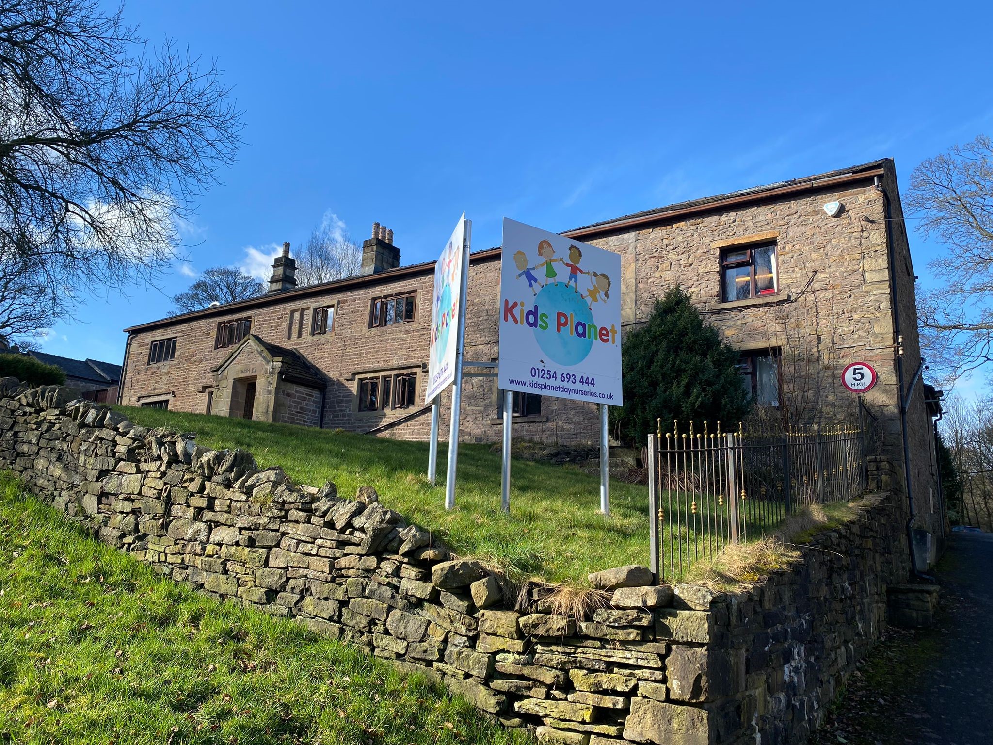 Kids Planet sign stands on a grassy slope in front of a stone building, advertising childcare; contact: 01254 693 444, www.kidsplanetdaynurseries.co.uk. Nearby wall shows a 5 MPH speed sign.