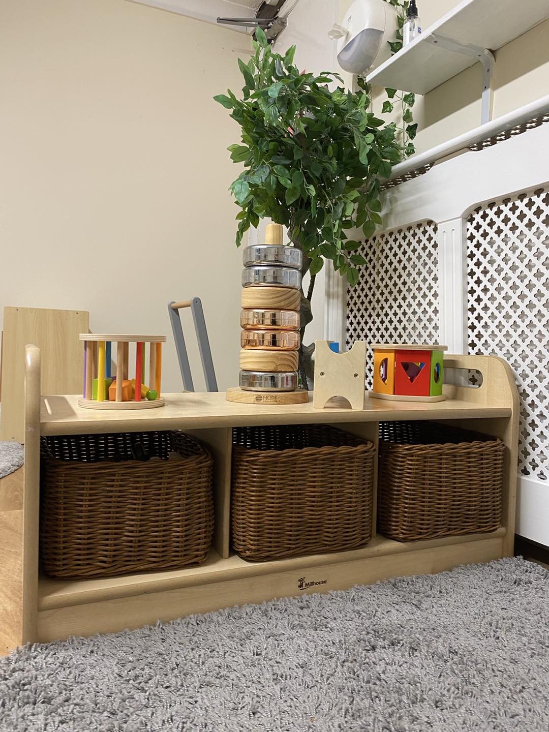 Wooden toy shelf holds three wicker baskets below and wooden toys—stacked rings, shape sorter, rattle—arranged on top, beside a potted plant and a white radiator cover in a carpeted corner.