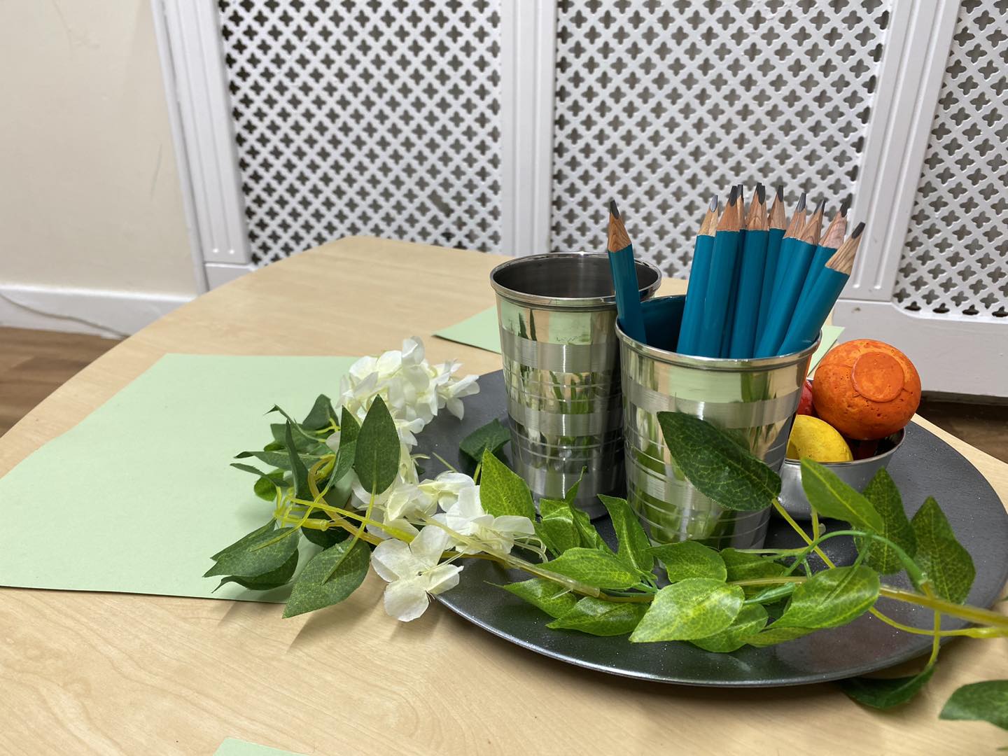 Metal cups hold sharpened teal pencils and small colored balls, arranged on a dark tray with artificial green leaves and pale-green papers atop a light wood table in front of a white lattice radiator cover.