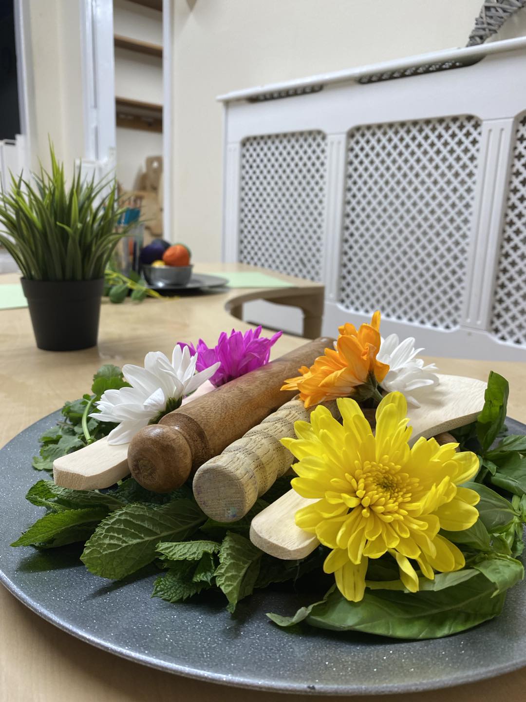 Wooden rolling pins and spatulas rest atop green leaves and multicolored flowers on a plate, set on a wooden table in a kitchen with a small plant and radiator cover.