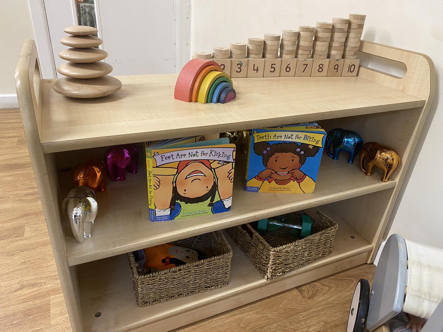 Wooden low shelf holds stacked wooden discs, a rainbow toy, and numbered blocks labeled 1 2 3 4 5 6 7 8 9 10; two board books titled "Feet Are Not for Kicking" and "Teeth Are Not for Biting." Lower shelves contain wicker baskets and metallic elephant toys against a pale wall.