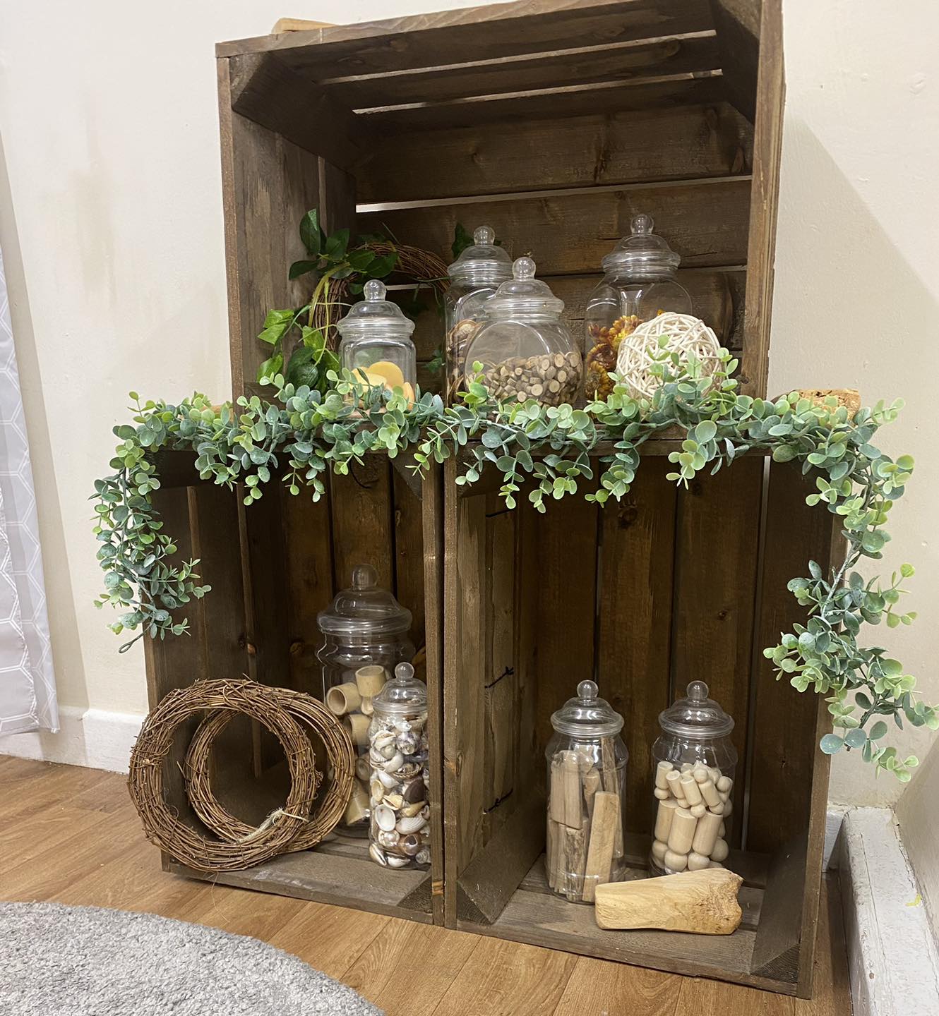 Wooden crates display glass apothecary jars and natural decorations; a trailing eucalyptus garland drapes across them on a hardwood floor against a pale wall in a cozy corner.