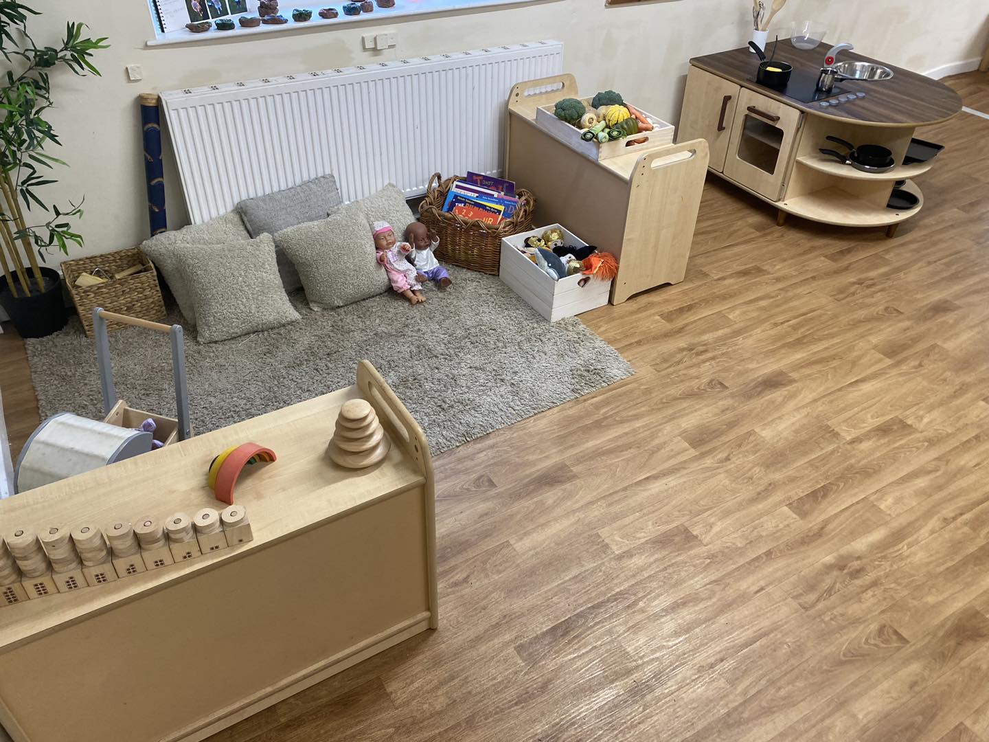 Dolls sit on a gray rug among three cushions, toys and baskets; nearby wooden crates hold toy food and books, and a small wooden play kitchen stands against a light wall.