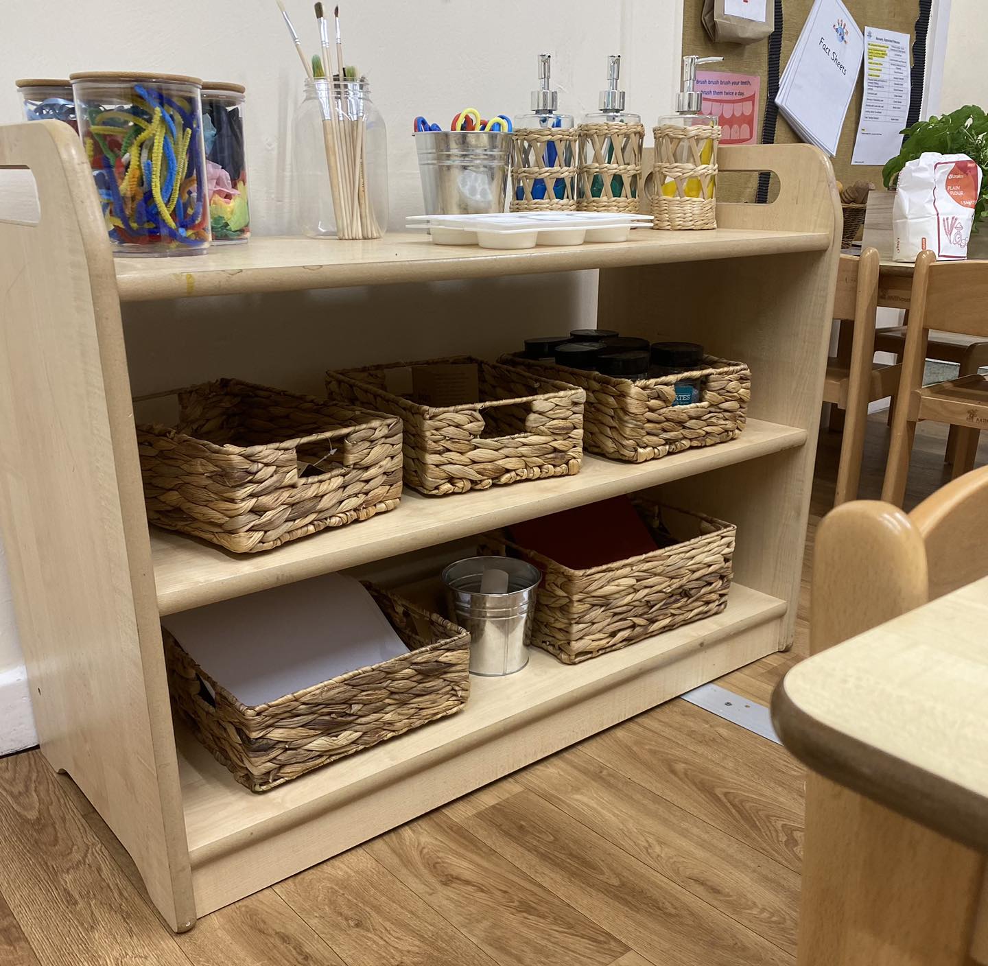 Wooden low shelf holds woven baskets, metal pails and jars of art supplies; paintbrushes, scissors and craft materials sit on top, in a small classroom with wooden chairs and floor.