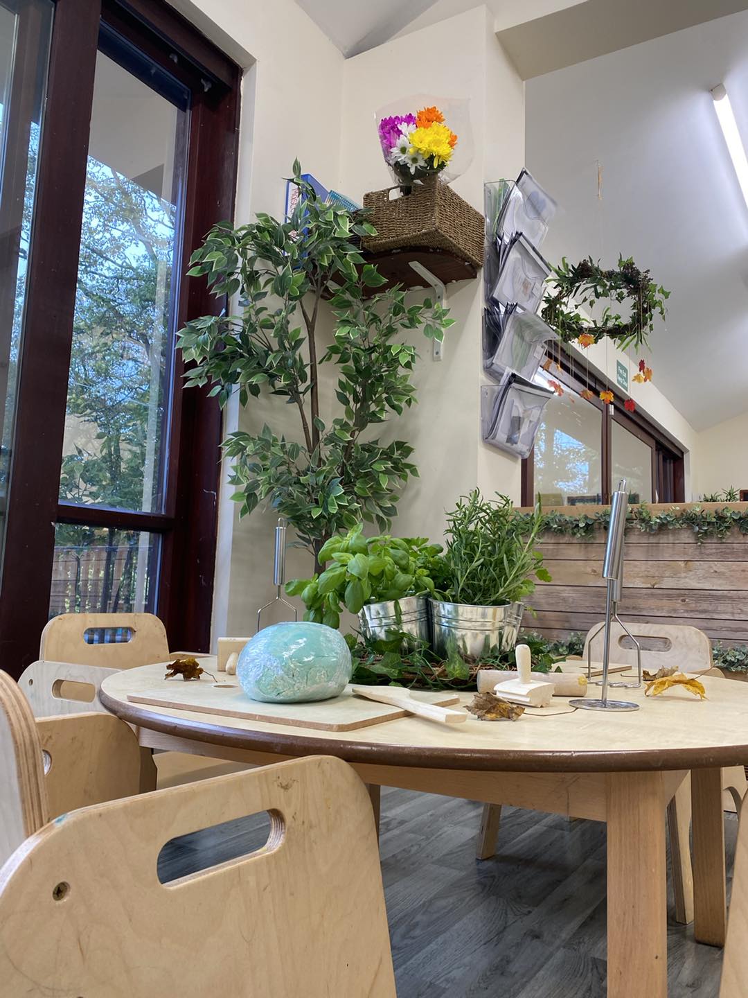 Round wooden table holds a turquoise ball of clay, rolling pins, metal herb pots and scattered leaves; surrounded by small wooden chairs, a tall faux tree, large windows and hanging wreaths.