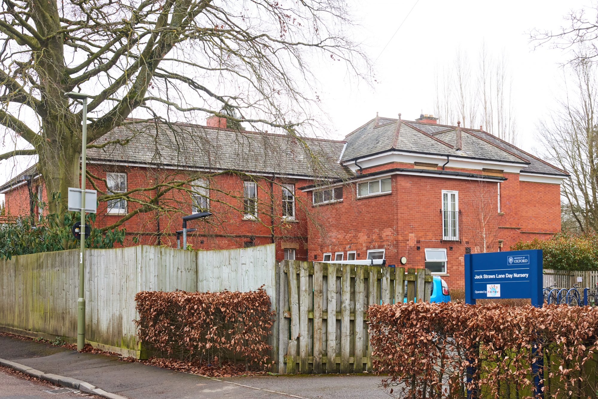 Red-brick nursery building stands behind a wooden fence; a blue sign reads "UNIVERSITY OF OXFORD" "Jack Straws Lane Day Nursery" (Operated by — text/logo not legible); leafless trees and hedges line the street.