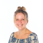 Woman smiling at camera, hair in a messy bun, wearing a blue-and-white patterned off-shoulder blouse, set against a bright white background.
