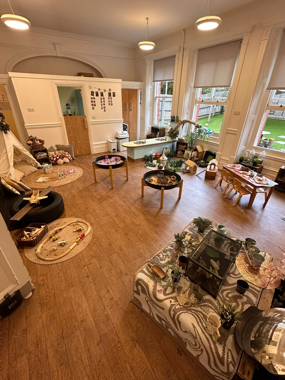 Children's playroom — multiple activity stations arranged with low tables, sensory trays, miniature houses and toys; cushions and craft materials set out for play; large windows look onto a fenced grassy yard.