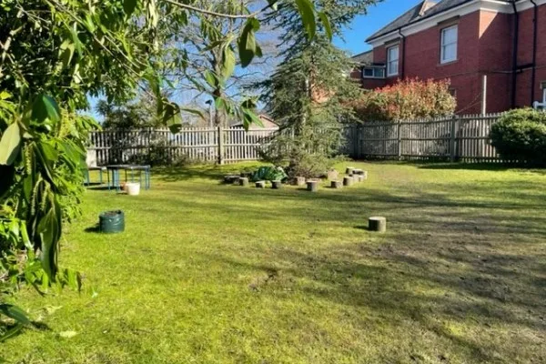 Backyard lawn contains scattered wooden stump seats, planters, a small round table and bins under a central tree, enclosed by a wooden fence beside red-brick house on a sunny day.