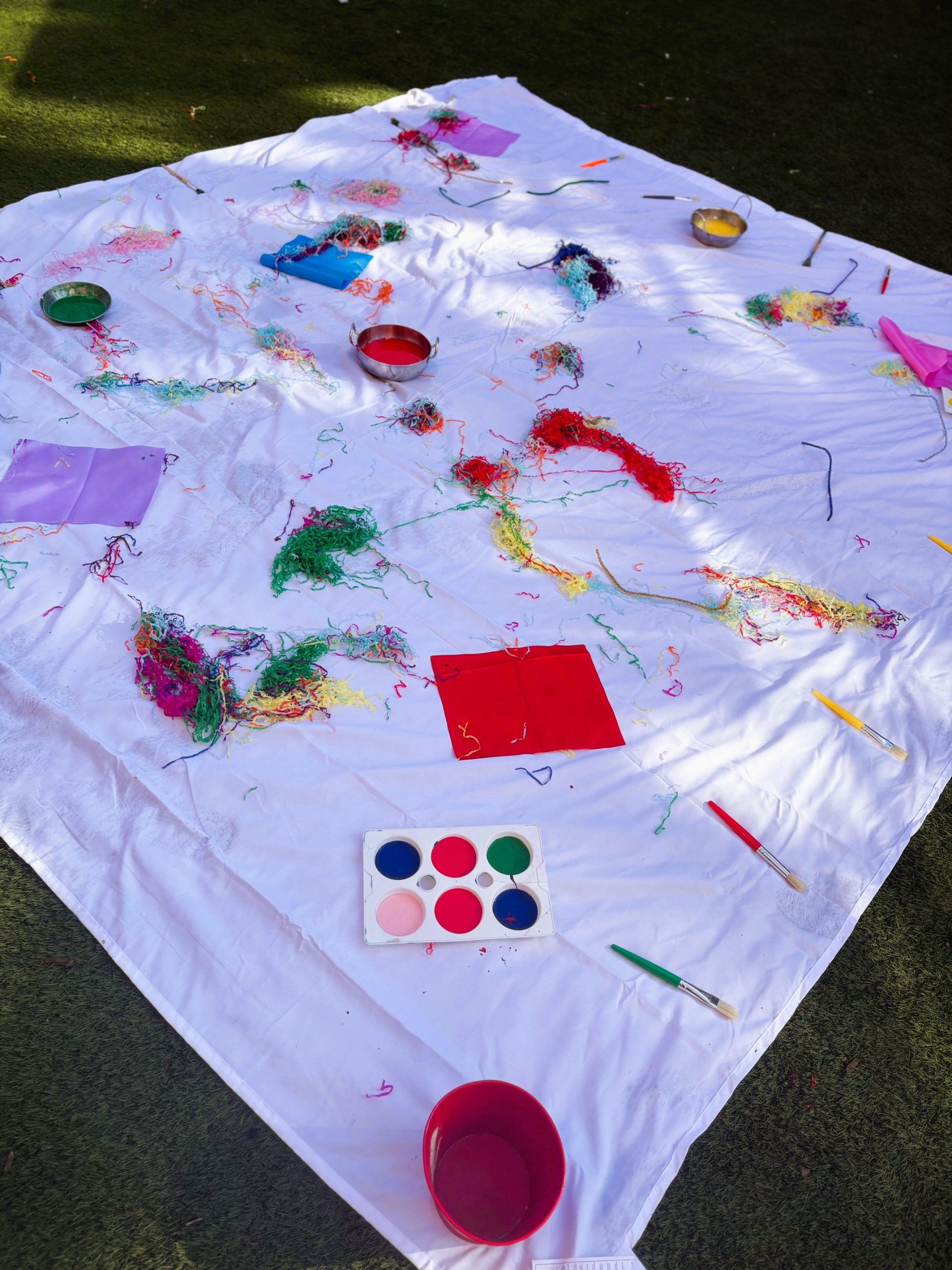 A large white sheet spread on grass, covered with a paint palette and bowls, brushes, shredded colorful yarn, and paper scraps; scattered art supplies indicate recent outdoor craft activity.