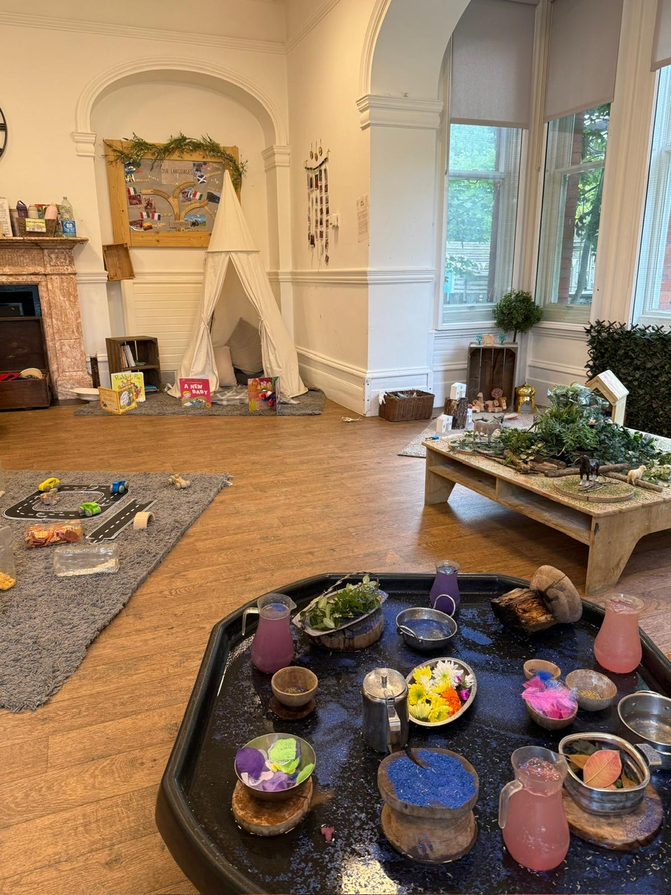 Low sensory tray holds cups, jars and pitchers with colored water, sand, flowers and leaves for play, in a children's classroom with a white teepee, rugs and large windows.
