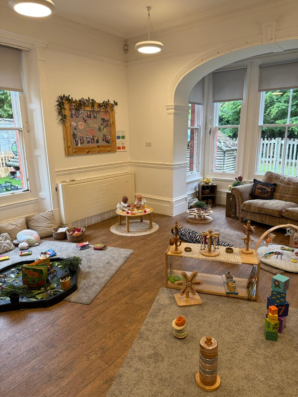 Children's wooden toys and soft play items are arranged on rugs and low tables, scattered across a sunlit bay-windowed playroom with a couch and a wall bulletin board.