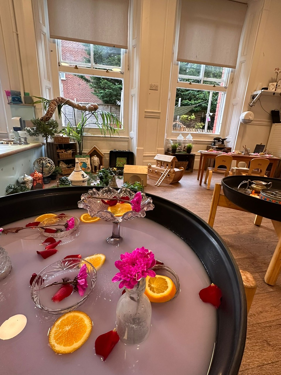 Black sensory tray holds milky pink water with floating orange slices, pink flowers and bowls; it sits in a sunlit children's playroom with wooden toys, small tables and large windows.