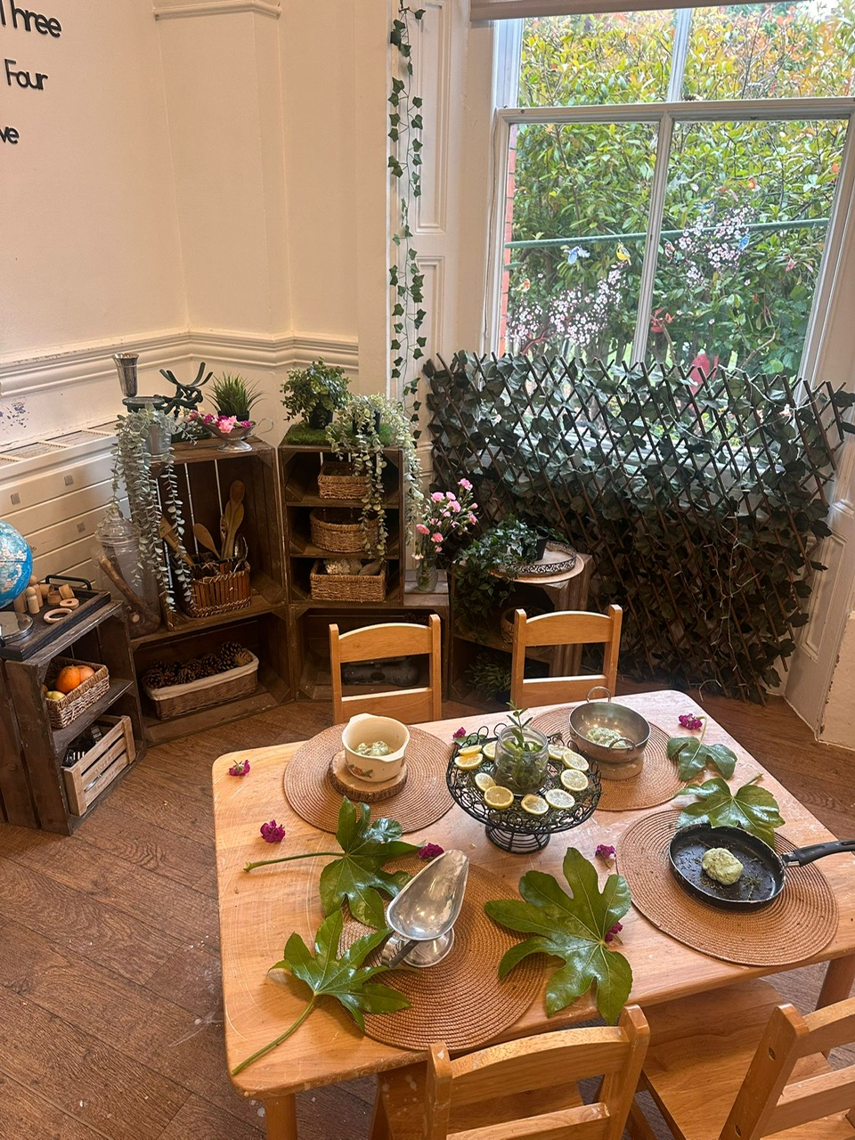 Wooden table arranged with placemats, lemons, pans and large leaves; three small chairs around it; in a cozy classroom corner with crates, potted plants and a leafy window. Text: "Three" "Four" "ve".