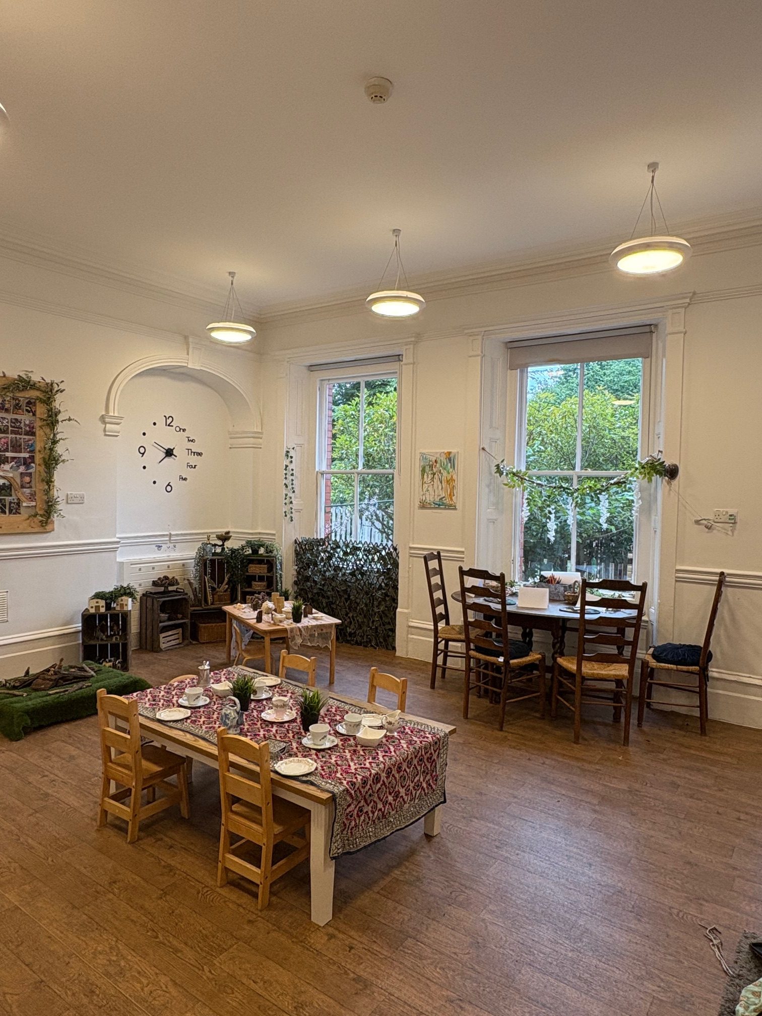 Small children's table arranged for a tea party with cups and plants; larger table and chairs nearby; tall windows show a garden; decorative wall clock and soft pendant lights.