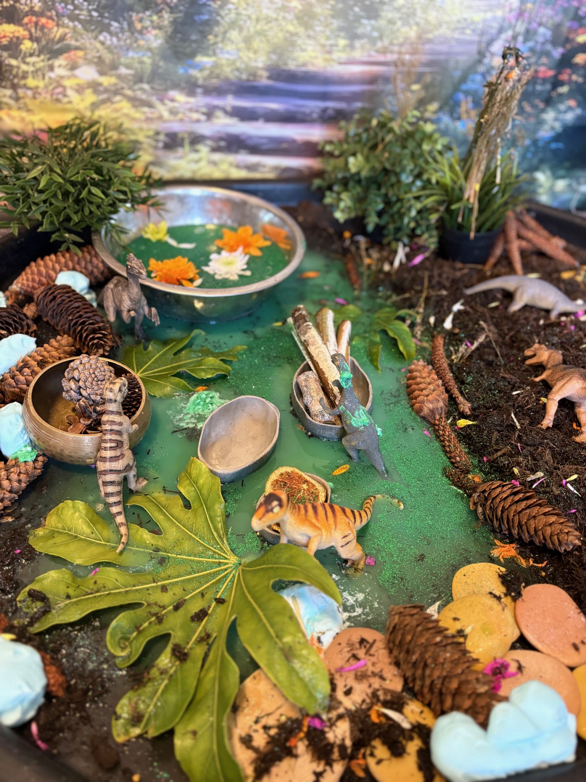 A small striped plastic dinosaur walks across a glittery green pond in a miniature forest diorama; toy reptiles, bowls with floating flowers, large leaves, pinecones, soil and stones surround it.