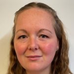 Close-up face of an adult looking directly at the camera, neutral expression, light freckles and flushed cheeks, subtle eyeliner, wavy hair pulled back, plain pale background.