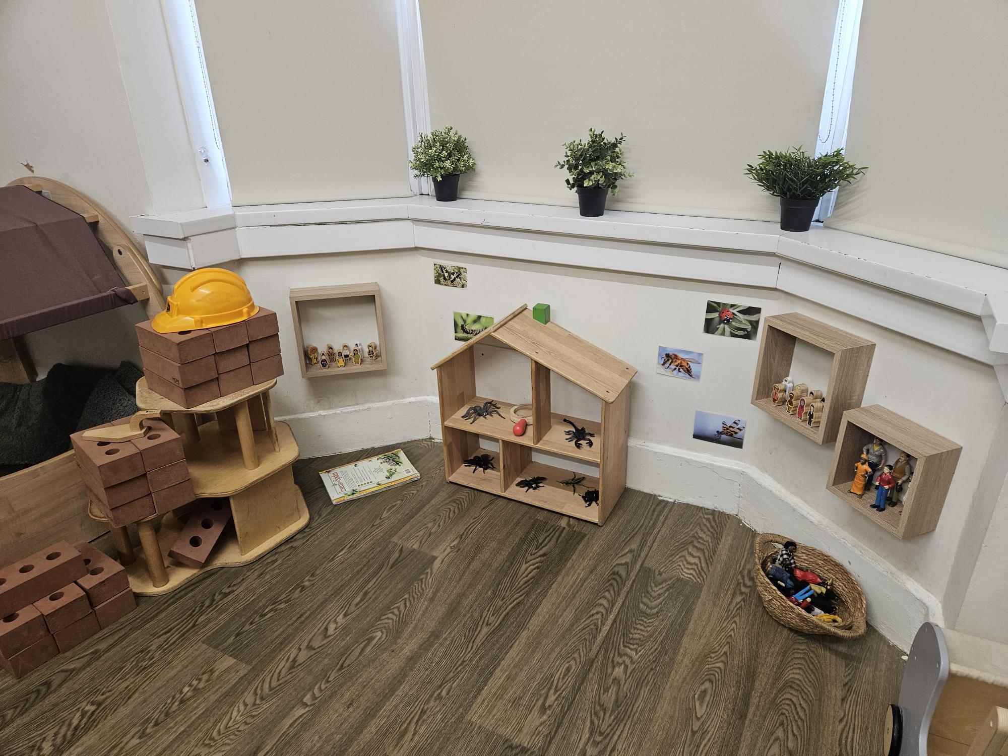 Wooden dollhouse holds plastic insects on shelves; wall cubbies display figurines; stacked toy bricks topped by a yellow hard hat sit on a stand; potted plants line the bay window.