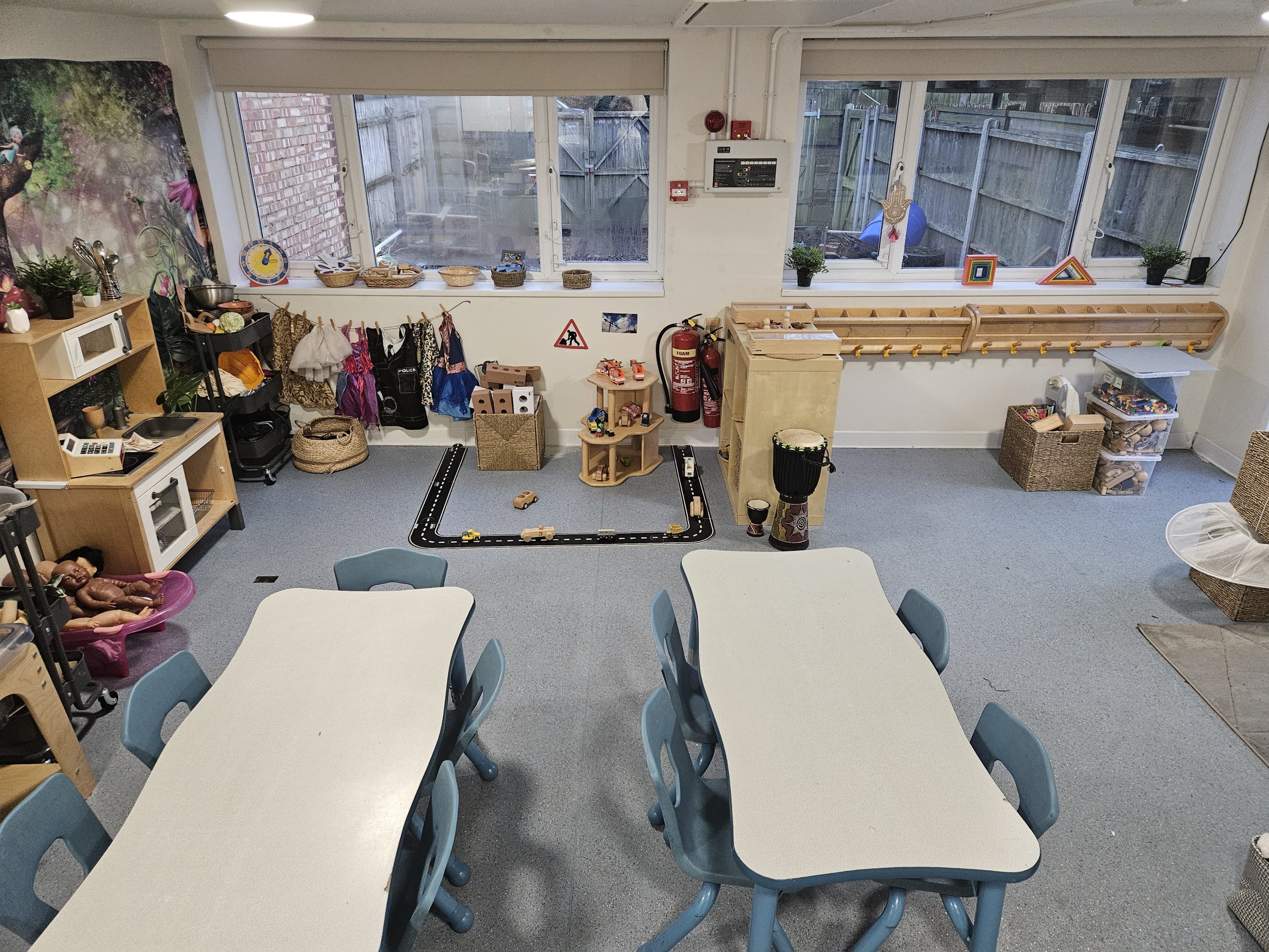 Two long white tables with blue chairs arranged for activities; a toy road with wooden cars on the floor, surrounded by dress-up costumes, baskets, a play kitchen, and windows.
