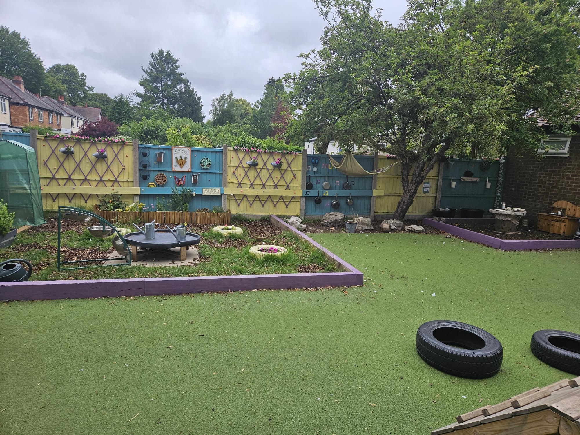 Colorful wooden fence panels adorned with hanging pots, signs, and toys stand behind a grassy play area; a leaning tree, raised planting beds, and two tires lie on artificial turf.