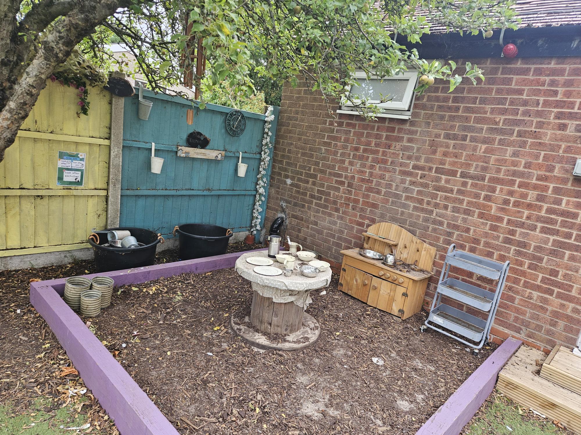 Wooden toy kitchen and spool table arranged with small plates and pots, sitting in a purple-edged mulch bed beside a brick wall and painted blue-yellow fence under an apple tree.