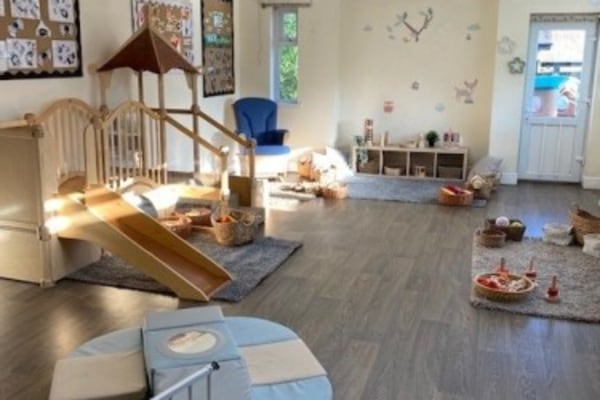 Wooden play structure with short slide and steps sits on a rug, surrounded by baskets of toys, soft seating, low shelves and wall decals in a sunlit preschool room.