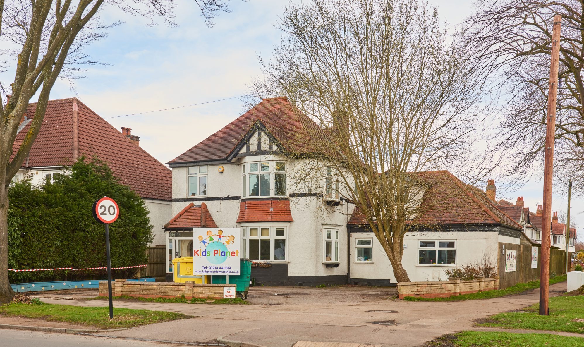 White two-story house converted into a childcare center stands behind a small forecourt, flanked by leafless trees and a 20 mph sign on a quiet suburban street.