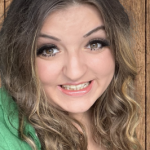 Woman with long wavy brown hair smiles broadly at camera, showing teeth; wearing a green top and eye makeup, posed against vertical wooden plank background.
