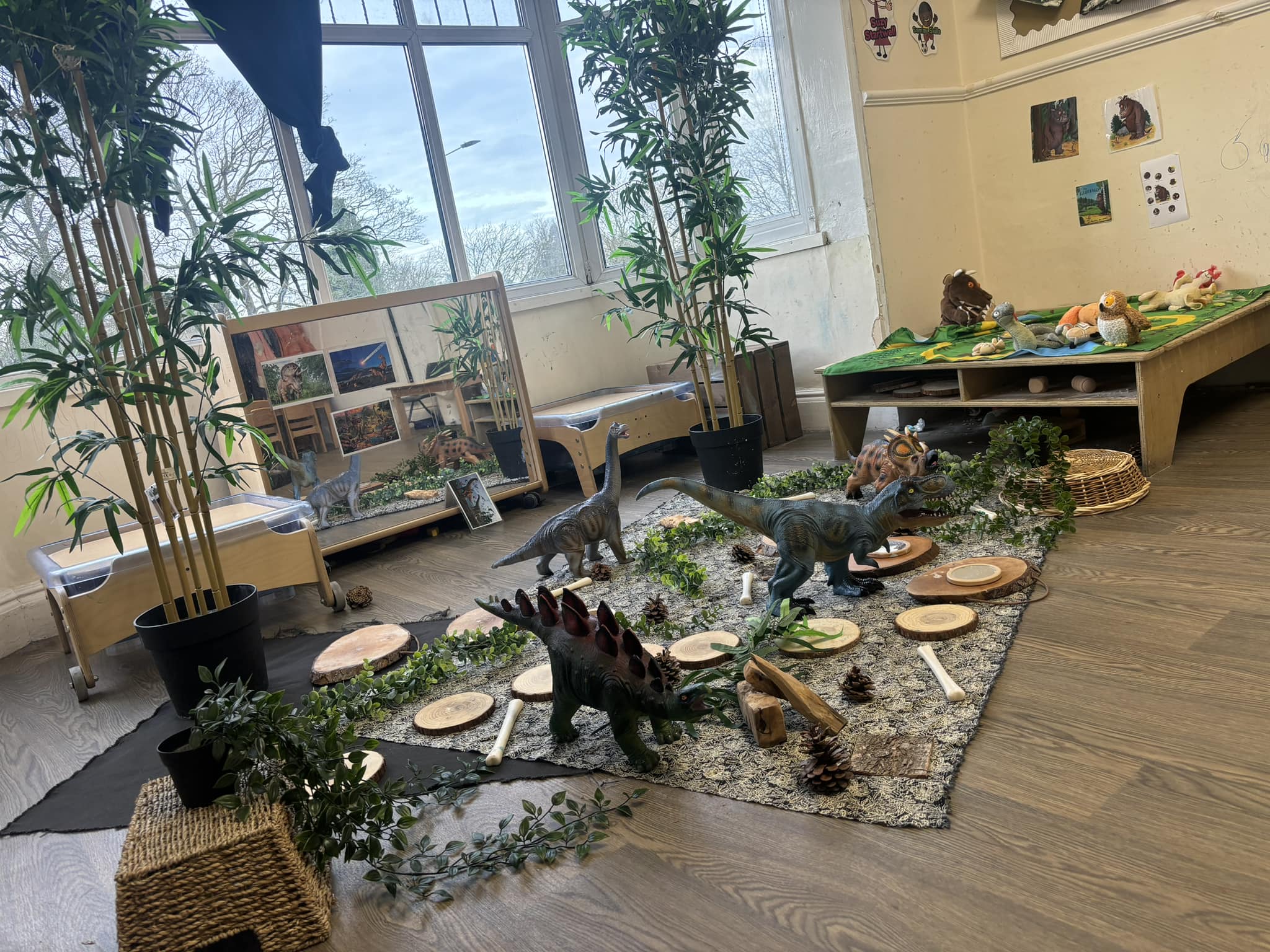 Toy dinosaurs are arranged among wooden slices, pinecones and faux plants on rugs, forming a play scene in a sunlit classroom with low shelves, stuffed animals, potted bamboo and window.