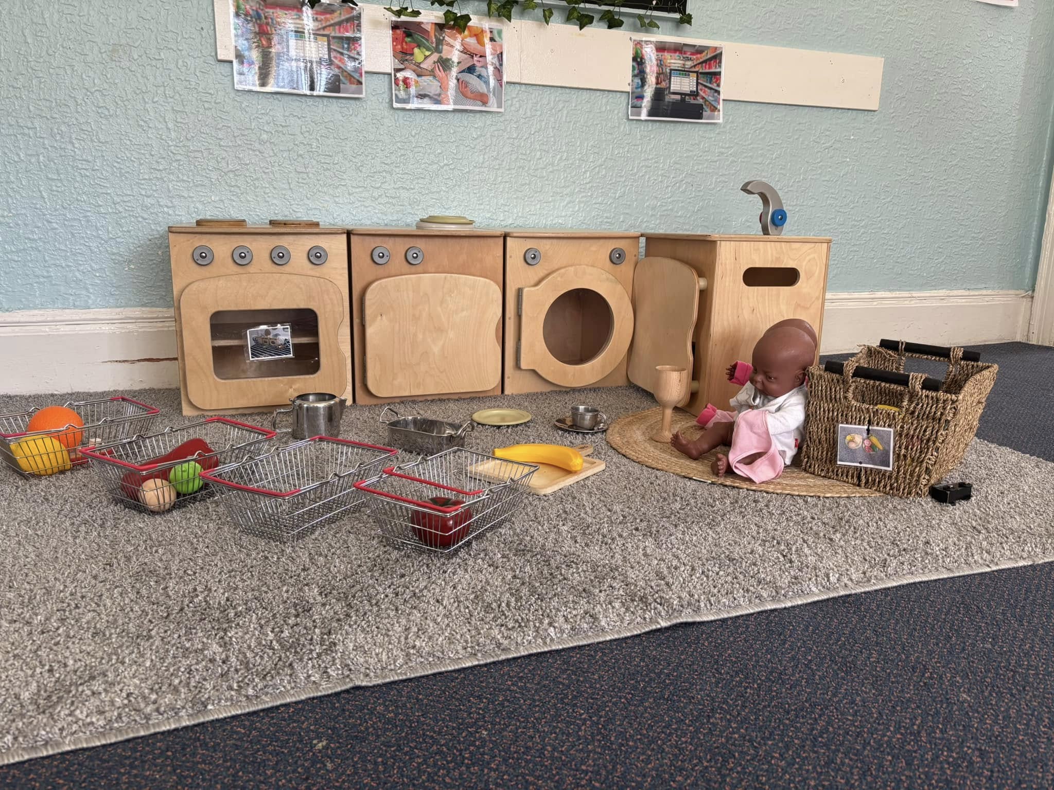 Wooden toy kitchen units sit along a light-blue wall, surrounded by metal shopping baskets of play food, with a seated baby doll on a mat in a carpeted play area.