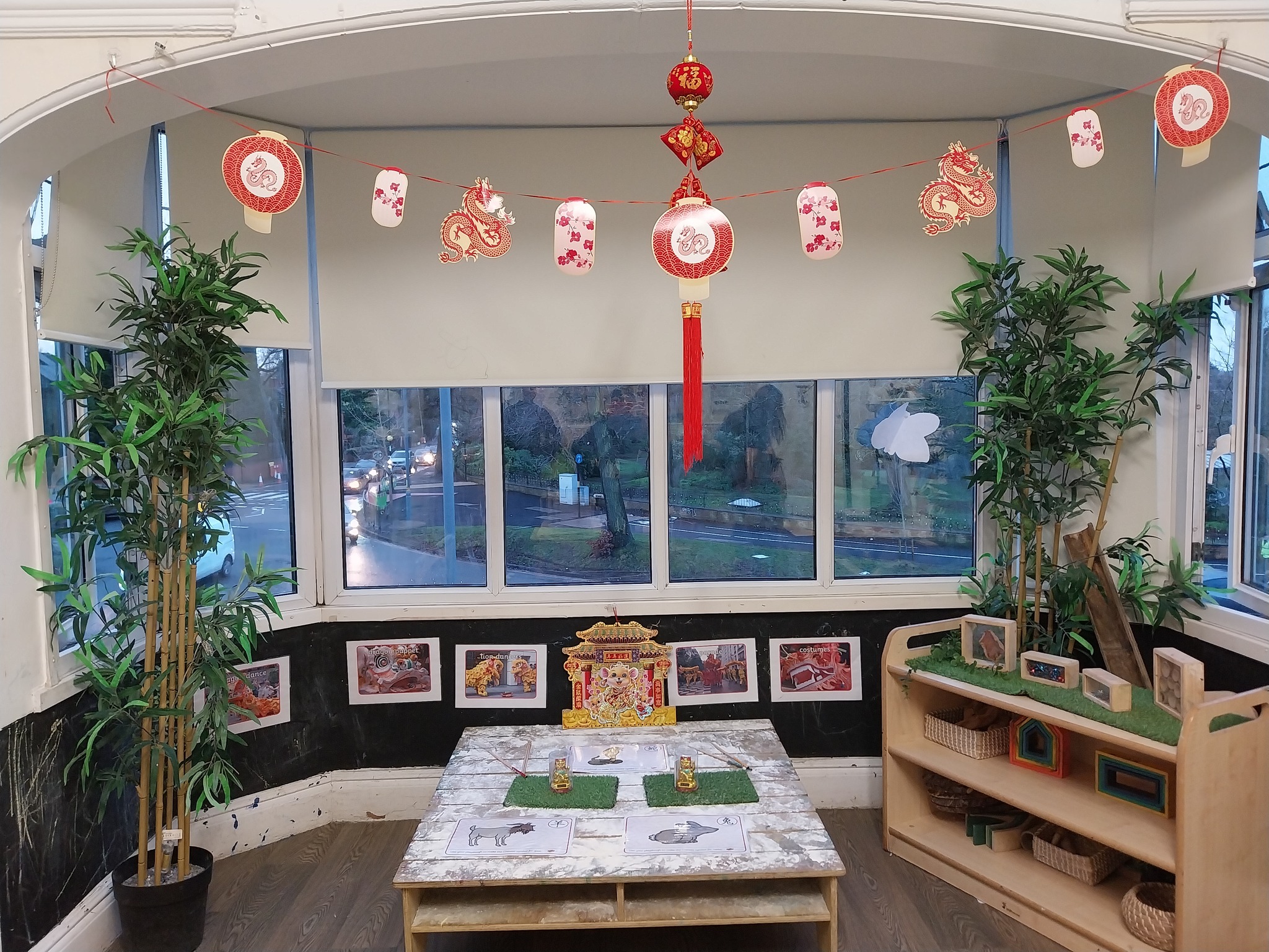 Red paper-lantern garland hangs across a bay window above a low craft table; two tall potted bamboo plants flank the scene in a classroom corner with shelves of educational toys.