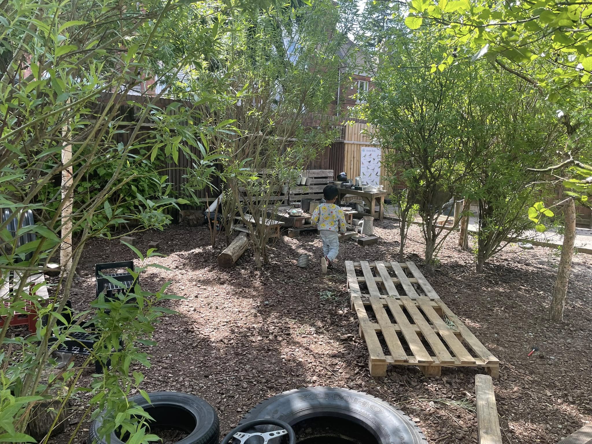 Child walking toward makeshift wooden table and pallets in a shaded, mulched backyard play area surrounded by small trees, old tires, logs, and scattered crates. Text: "KUMHO" (on tire).