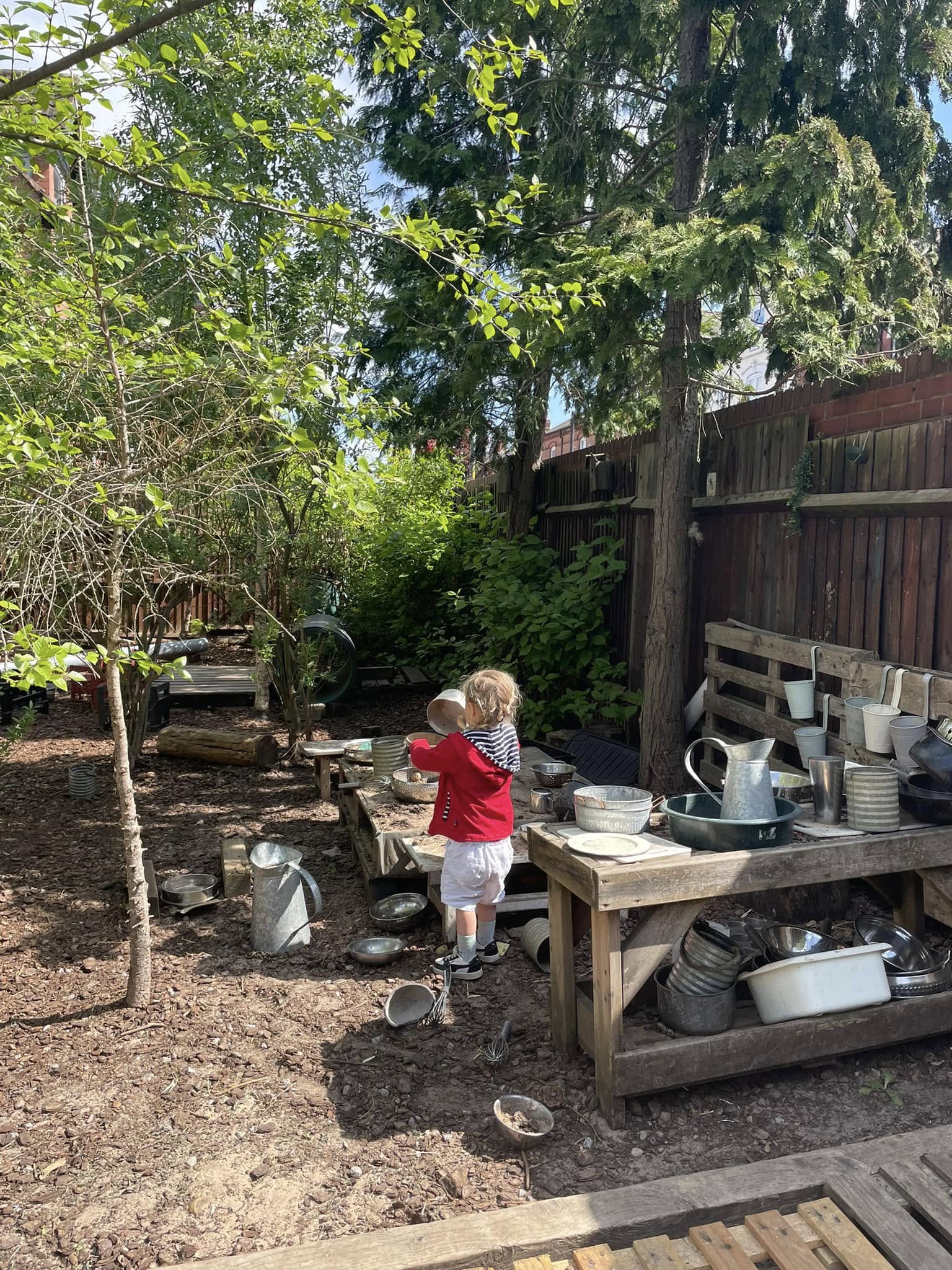 Child in a red jacket pours from a metal bowl at a wooden bench amid scattered pots and pitchers in a shaded backyard garden with trees, mulch and wooden fence.
