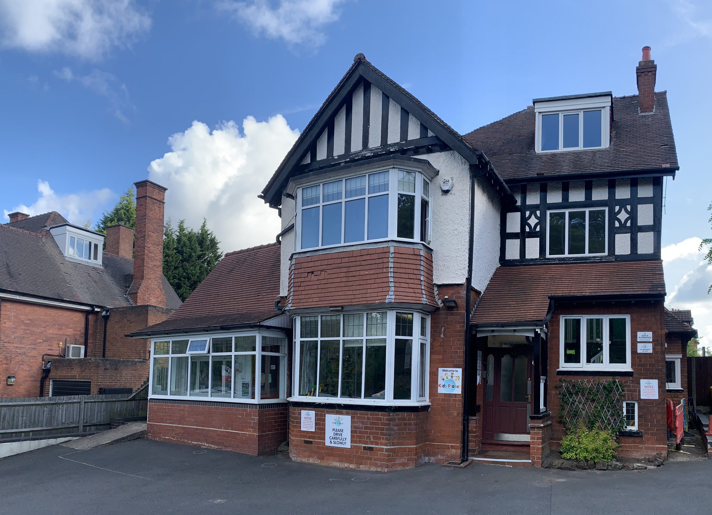 A two-story Tudor-style brick-and-timber house displays childcare signage, standing by a paved forecourt under a blue sky with scattered clouds.
