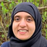 Person wearing a black hijab smiles directly at the camera; close-up of the face against blurred green shrubbery outdoors.