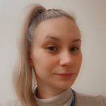 Woman with blonde ponytail — looking into camera with a slight smile — plain indoor background, wearing a light turtleneck and a blue lanyard reading 