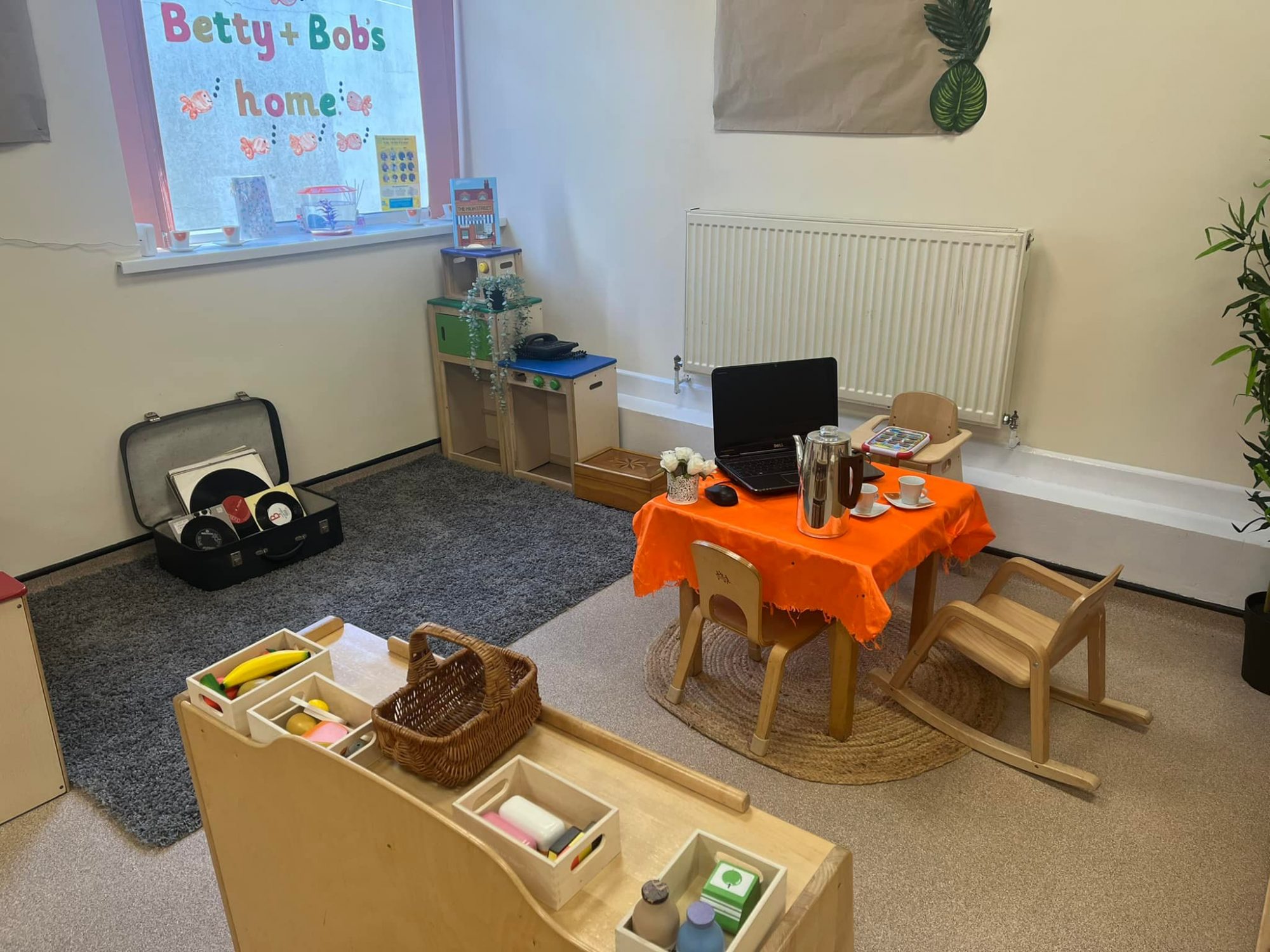 Small child-sized table holding an open laptop, metal coffee pot, toy teacups and a vase (set up for pretend tea); placed on a round rug in a carpeted preschool corner with shelves and toys. Text: "Betty + Bob's home"