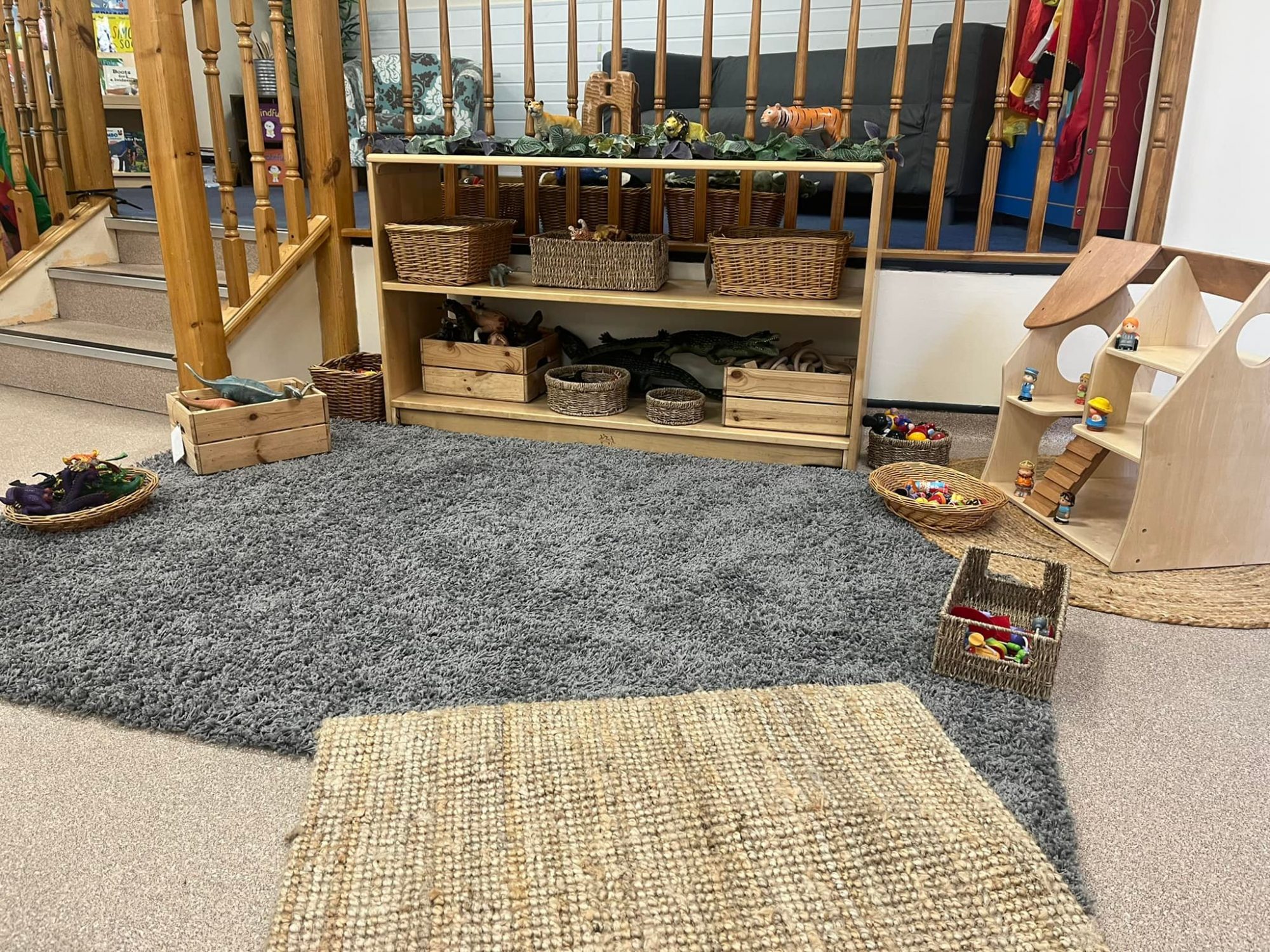 Low wooden shelf holds wicker baskets and toy animals, arranged against wooden railings beside a gray rug; nearby wooden dollhouse, baskets of toys, carpeted steps and seating area.