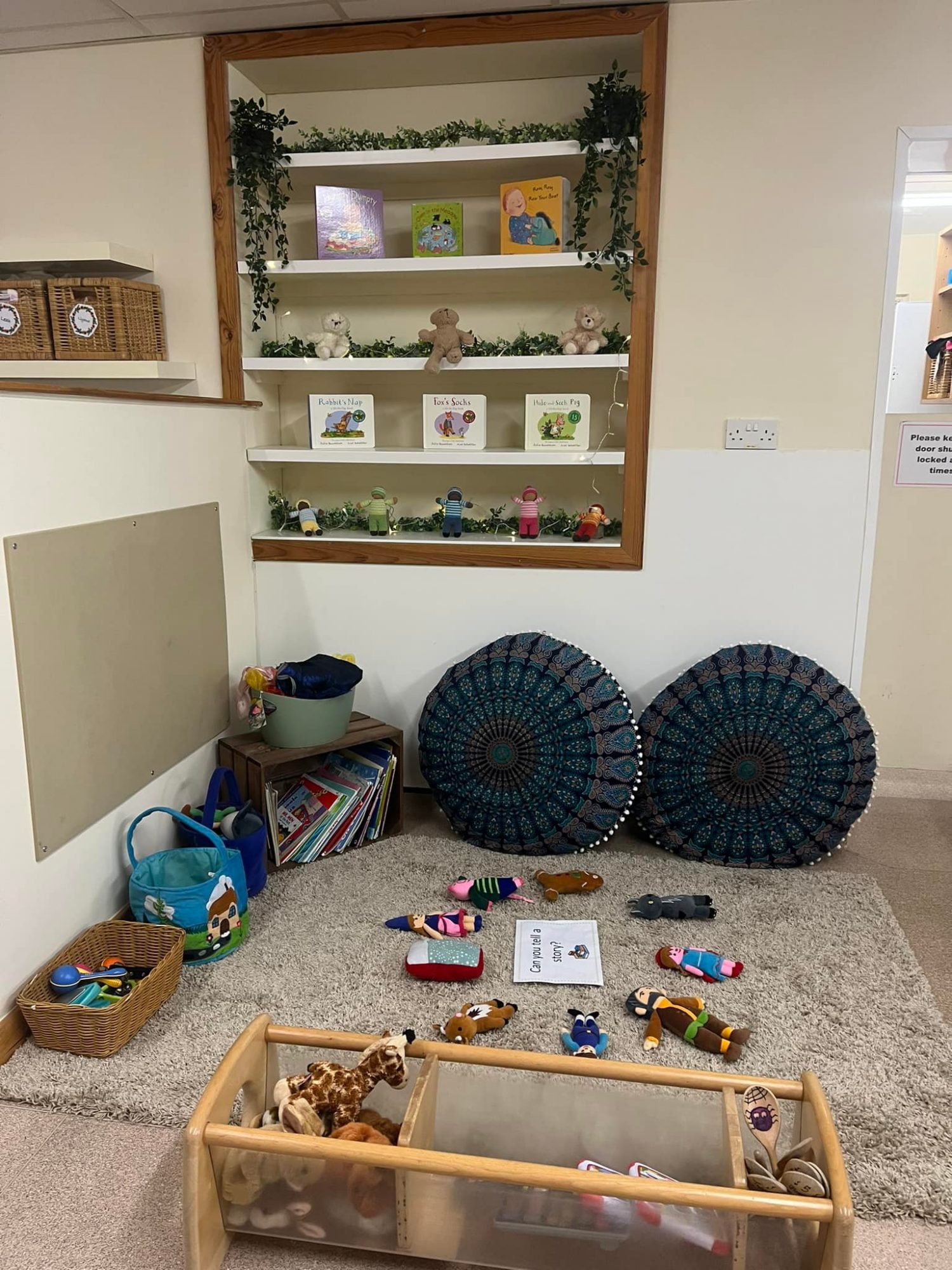 Children’s reading corner: stuffed animals and cloth dolls scattered on a rug, two round patterned cushions propped, wooden shelves displaying picture books and small toys in a classroom nook.