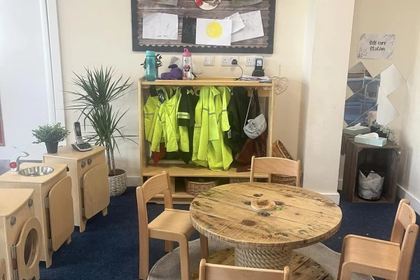 Wooden round children's table with small chairs sits before a low cubby of fluorescent safety jackets and a toy kitchen; potted plants and carpet define a preschool play corner.