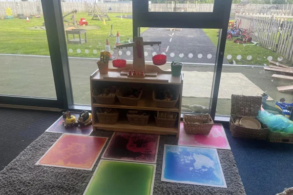 Wooden low shelf topped by a toy balance scale with red pans holding items; open baskets of toys in shelves; colorful square mats on carpet; glass doors reveal outdoor playground.