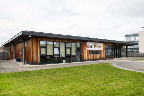 One-story wood-clad building displays the sign "Kids Planet", set beside a paved entrance and a green lawn, with an overcast sky and modern buildings visible in the background.