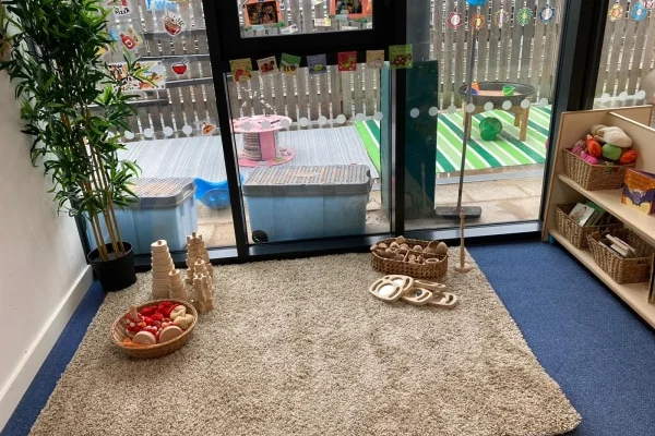 Wicker baskets of wooden play food sit arranged on a beige rug beside a bookshelf in a childcare playroom with sliding glass doors to a fenced outdoor play area.