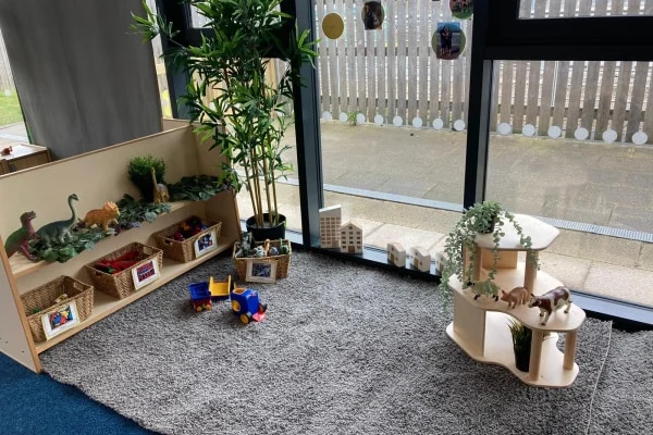 Low wooden shelves and a curved stand display toy animals and wicker baskets; a small plastic truck rests on a gray rug by a potted plant beside floor-to-ceiling windows overlooking a patio.