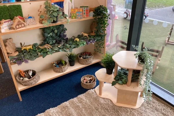 Wooden low shelf displays toy animals, miniature houses and woven baskets with plastic dinosaurs and plants; small wooden play table holds potted greenery in carpeted classroom corner by glass wall.