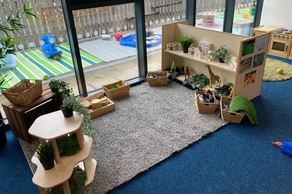 Low wooden shelf holds toy animals, baskets, and potted plants on a gray rug; play materials spread around in a bright preschool room beside glass doors to a toy-filled deck.