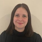 Adult woman with shoulder-length brown hair smiles at the camera, wearing a black turtleneck and dark jacket, posed against a plain off-white wall in neutral indoor lighting.
